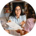 A circular image of a woman wearing glasses, talking with another person about a paper she's referring to