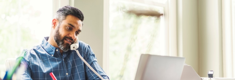 Man sitting at a home office, talking on the phone, writing on a notepad, and looking at a laptop screen