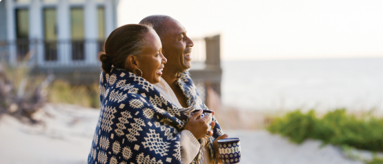 An older man and woman, wrapped in a blanket, on a beach, overlooking the ocean smiling