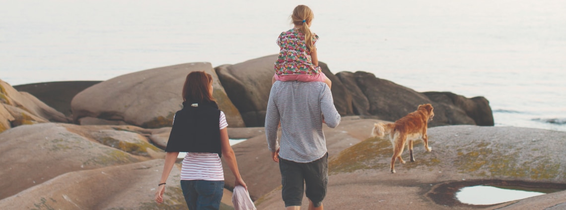 dad walking with daughter on shoulders with mother following and dog ahead on rocks near a body of water